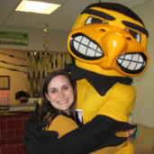 Jessi Beck wearing gold and smiling while hugging Herky, the black and gold hawkeye mascot