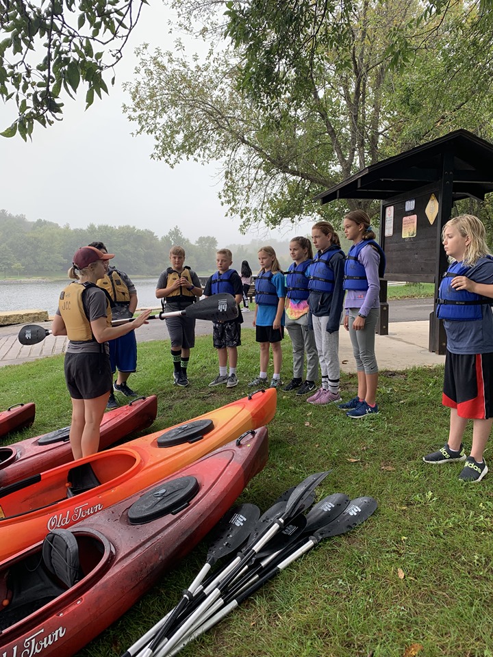 A naturalist talks to a group of middle school students wearing life jackets and standing by a row of kayaks.