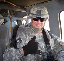 Alan Kellogg sitting in a aircraft smiling wearing his Army gear with helmet and sunglasses giving a thumbs up
