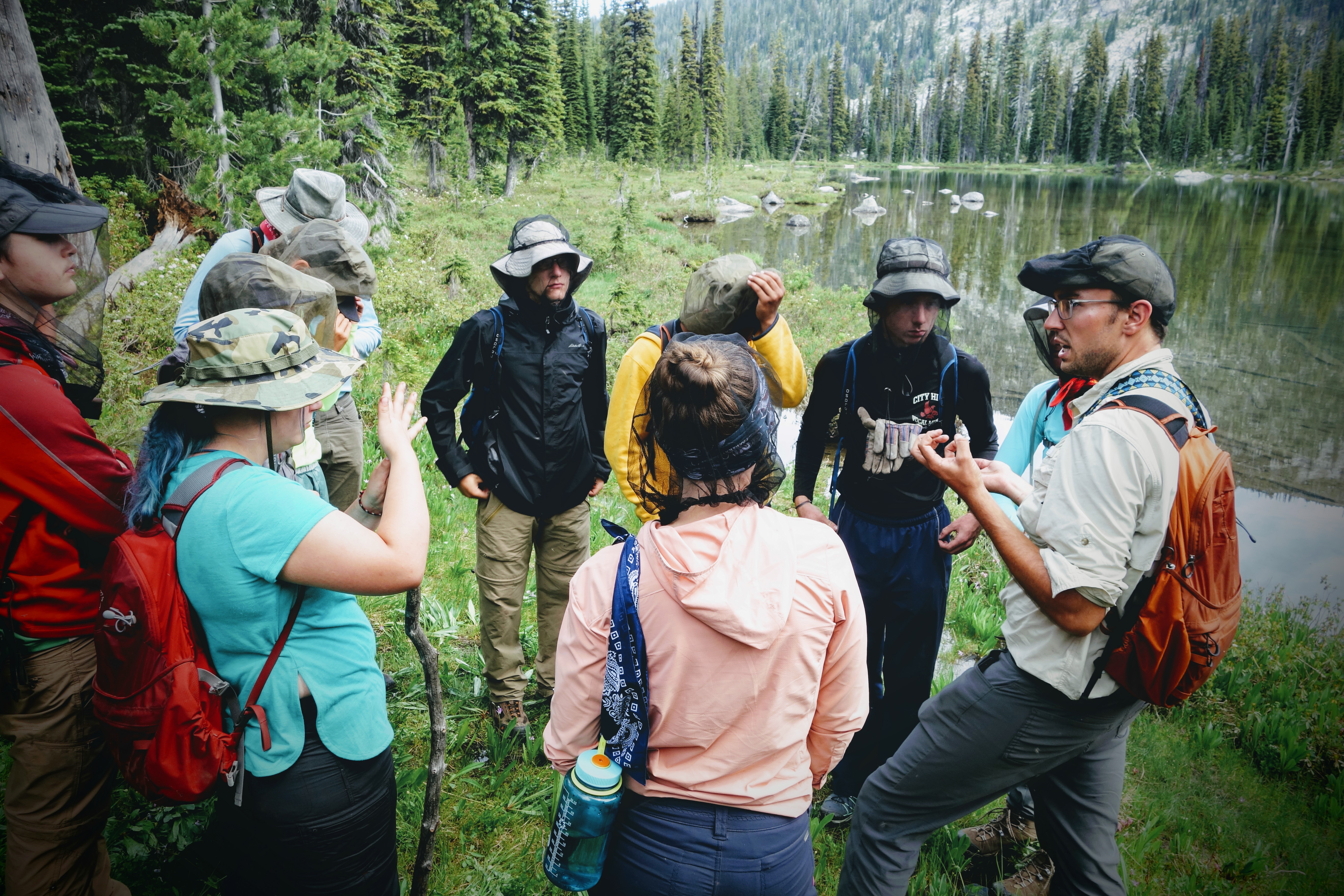 A trip leader speaks near a lake while a group of high school students is circled around him listening.