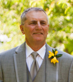 Joseph Gasperetti smiling at a wedding wearing a gray tux with a yellow flower pinned to the left coller
