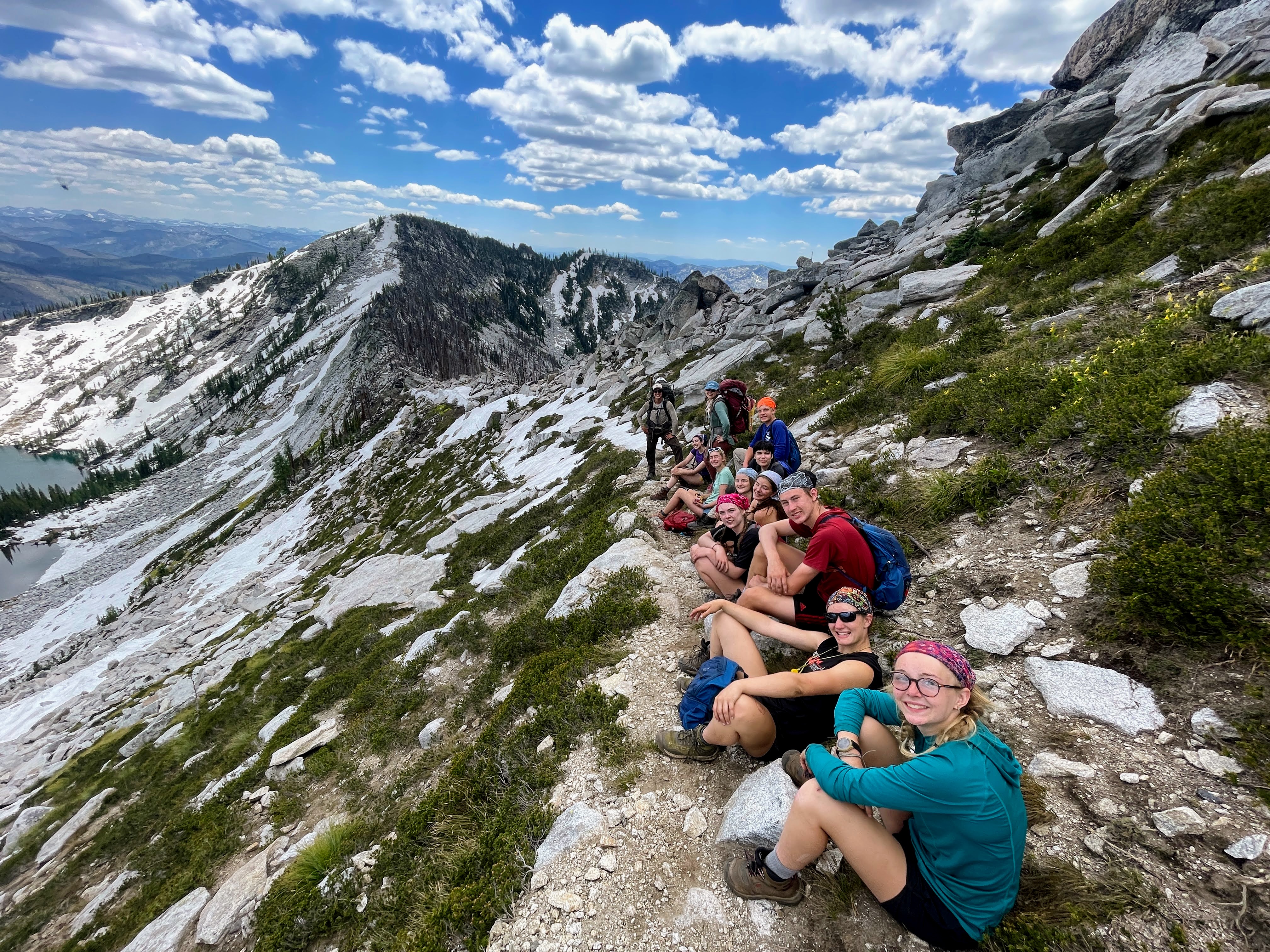 A group of teenagers sit in a line along a trail that ascends up a mountain. Mountain peaks fill the backdrop.