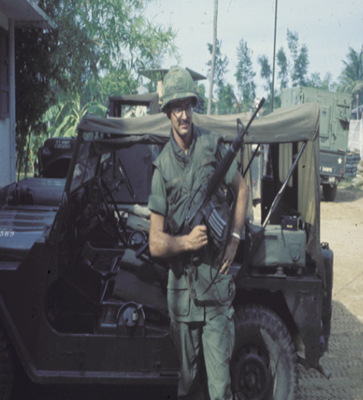 Palmer Holden standing next to an Army jeep holding a rifle in his field gear
